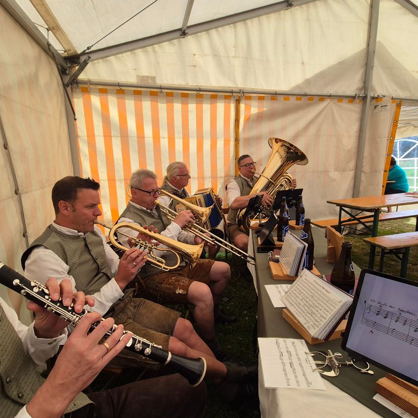 Eine Gruppe von Musikern in traditioneller Kleidung spielt Blechblasinstrumente in einem Zelt. Ein Monitor mit Notenblättern steht auf dem Tisch vor ihnen.