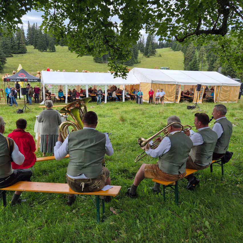 Eine Gruppe von Musikern in traditioneller Kleidung sitzt auf Bänken in einem Grasfeld und spielt Instrumente unter einem Zeltdach, mit Menschen und einer Bergkulisse im Hintergrund.