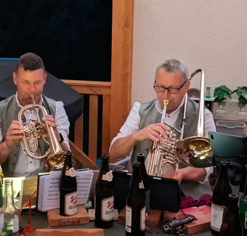 Zwei Männer spielen Trompeten an einem Tisch mit Bierflaschen und Notenblättern. Ein Mann trägt eine Brille und eine Weste.