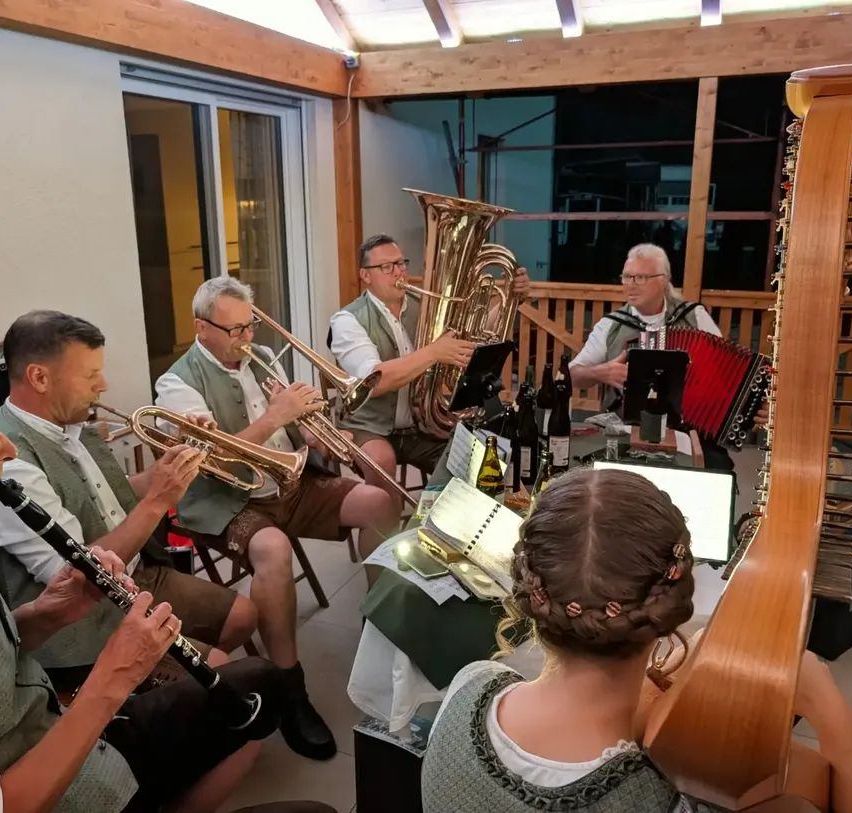 Eine Gruppe von Musikern spielt in einem Raum Instrumente, darunter Tuba, Trompete und Akkordeon. Sie tragen traditionelle Kleidung.