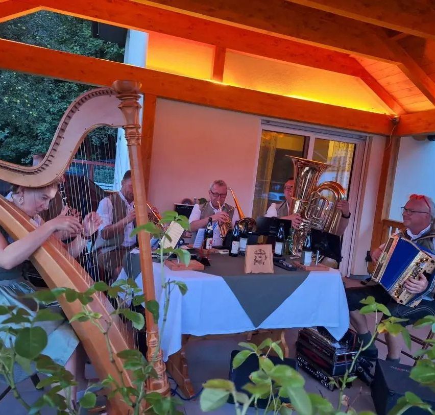 Eine Gruppe von Musikern spielt auf einer Terrasse mit Harfe, Tuba und anderen Instrumenten. Die Umgebung ist im Freien mit einem Holzdach.