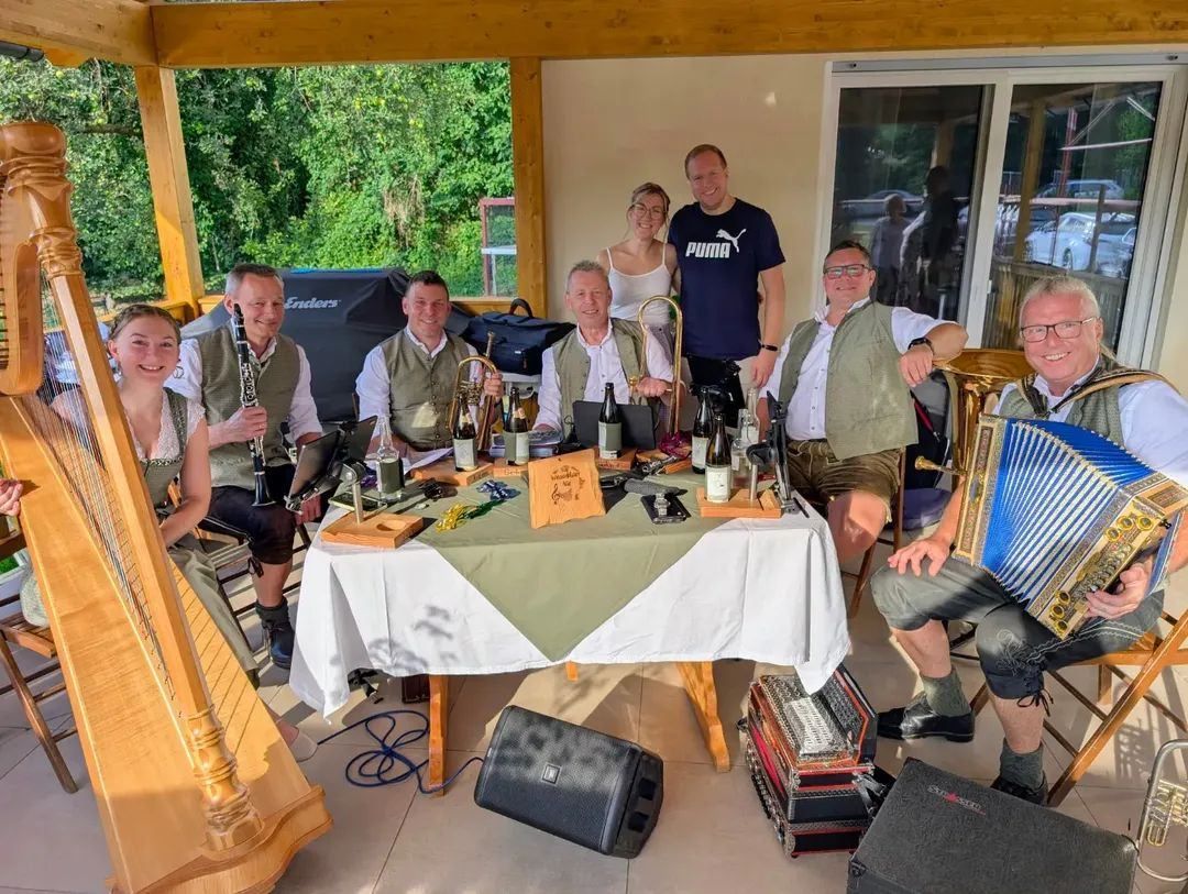 Eine Gruppe von Musikern, darunter eine Frau, die Harfe spielt, versammelt sich um einen Tisch mit Flaschen und Musikinstrumenten. Sie sind in traditioneller Kleidung gekleidet.
