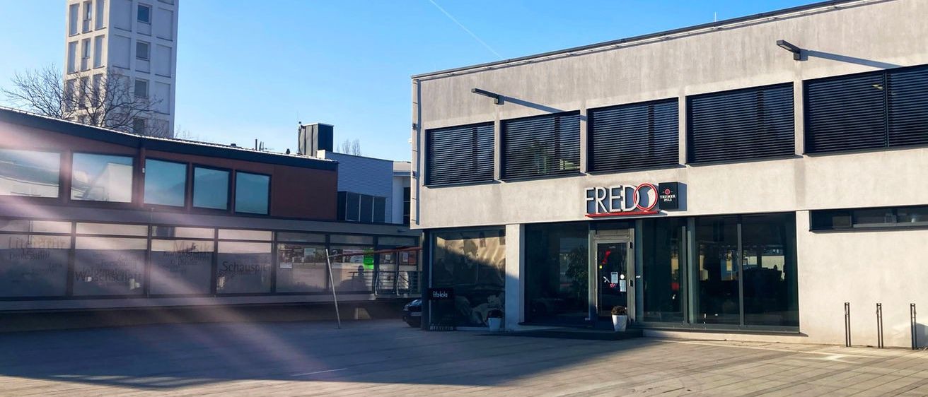 Ein modernes Gebäude mit dem Namen Fredo steht neben einem Uhrturm unter einem klaren blauen Himmel. Der Bereich davor ist leer mit einem Holzboden.