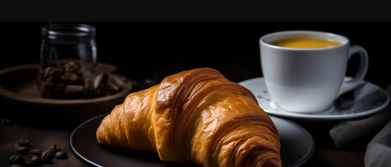 Eine Nahaufnahme eines goldbraunen Croissants mit glänzender, blättriger Textur, auf einer schwarzen Oberfläche mit einer weißen Tasse Kaffee im Hintergrund.