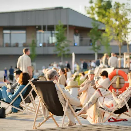 Eine Gruppe von Menschen sitzt auf Stühlen auf einer Holzterrasse vor einem Gebäude. Einige tragen Hüte und Brillen. Ein Rettungsring hängt an der Brüstung.