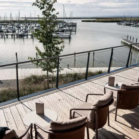 Ein Blick von einem Balkon mit einer Brüstung auf eine Marina mit angedockten Booten und einem Baum im Vordergrund. Die Terrasse ist mit gepolsterten Stühlen und Tischen möbliert.