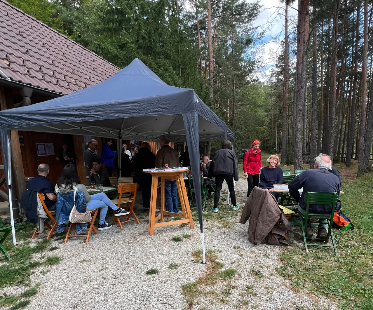 Eine Gruppe von Menschen versammelt sich unter einem Zelt in einer Waldumgebung, einige sitzen um Tische herum, andere stehen und unterhalten sich. Das Zelt ist blau und die Umgebung ist üppig mit Bäumen.