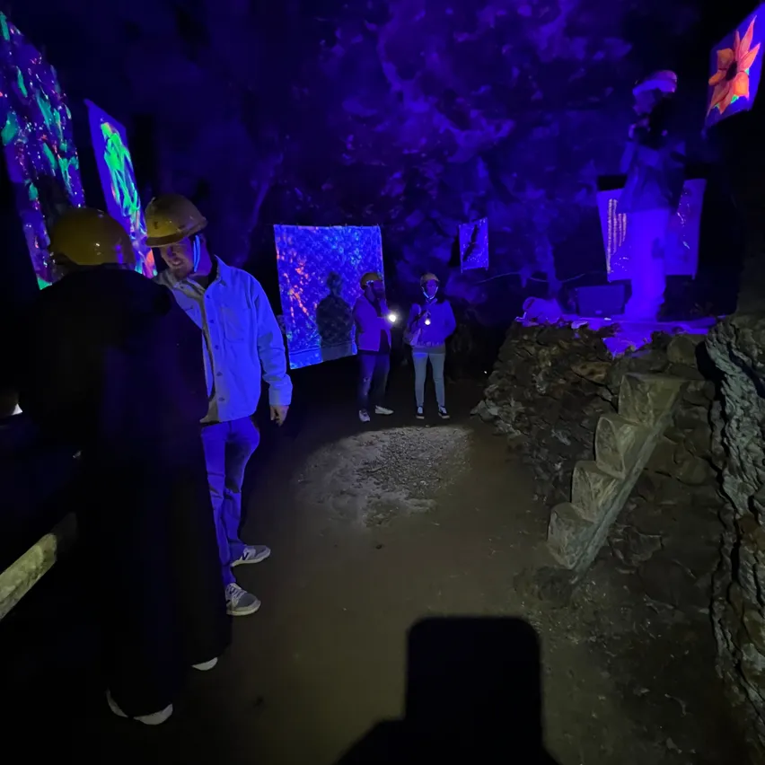 Eine Gruppe von Menschen in einer dunklen Höhle mit leuchtenden blauen Lichtern, die Helme tragen und Taschenlampen halten. Es gibt Steinstufen und Wände mit leuchtender Kunst.