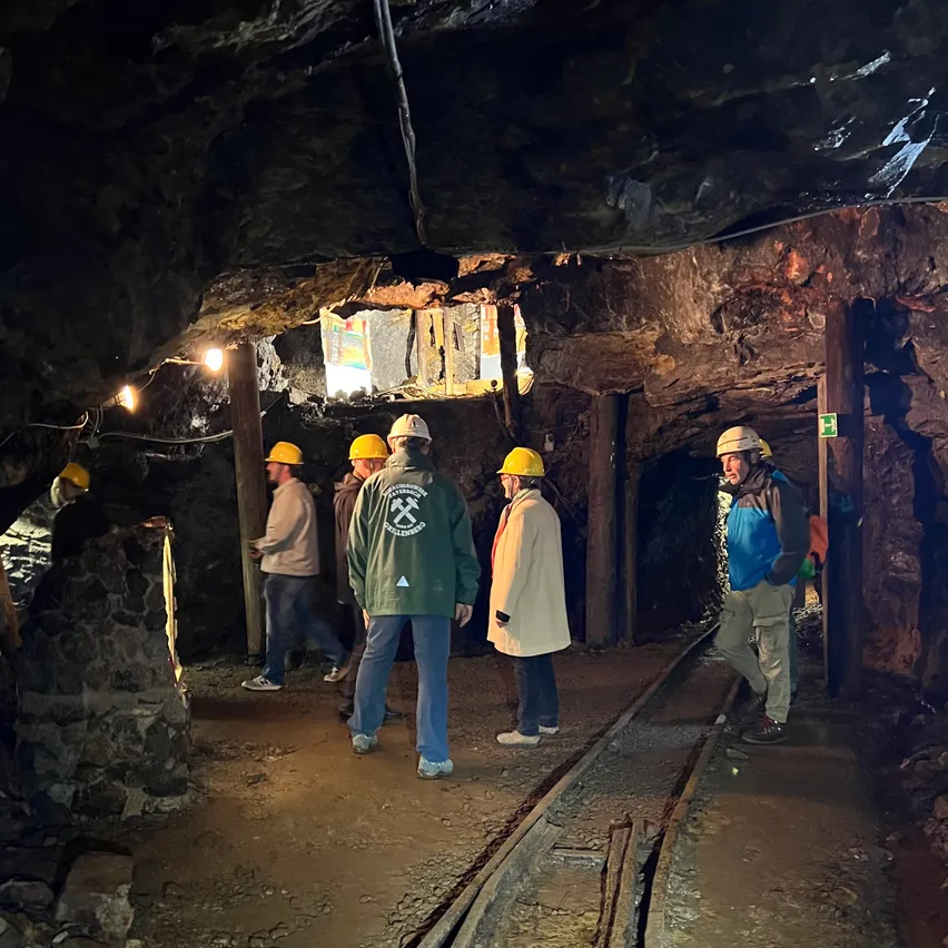 Eine Gruppe von Menschen in Schutzhelmen geht durch einen Minentunnel mit Gleisen und einer Lampe an der Decke.