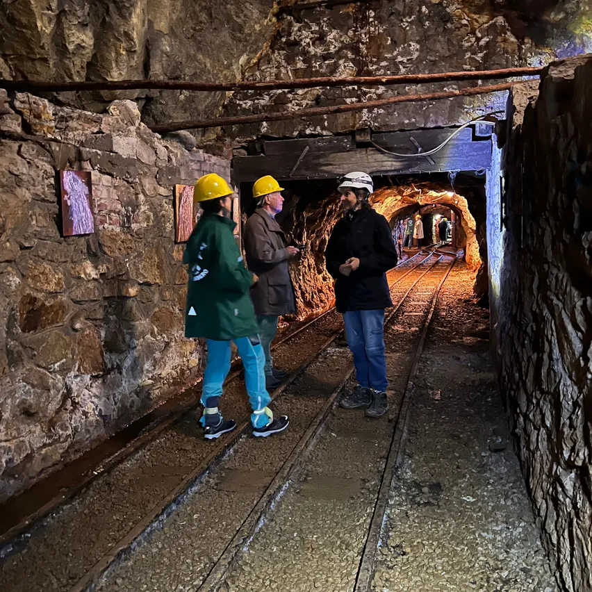 Drei Personen in Schutzhelmen stehen in einem Tunnel mit Steinwänden und einer Eisenbahnschiene. Der Tunnel ist schwach beleuchtet.