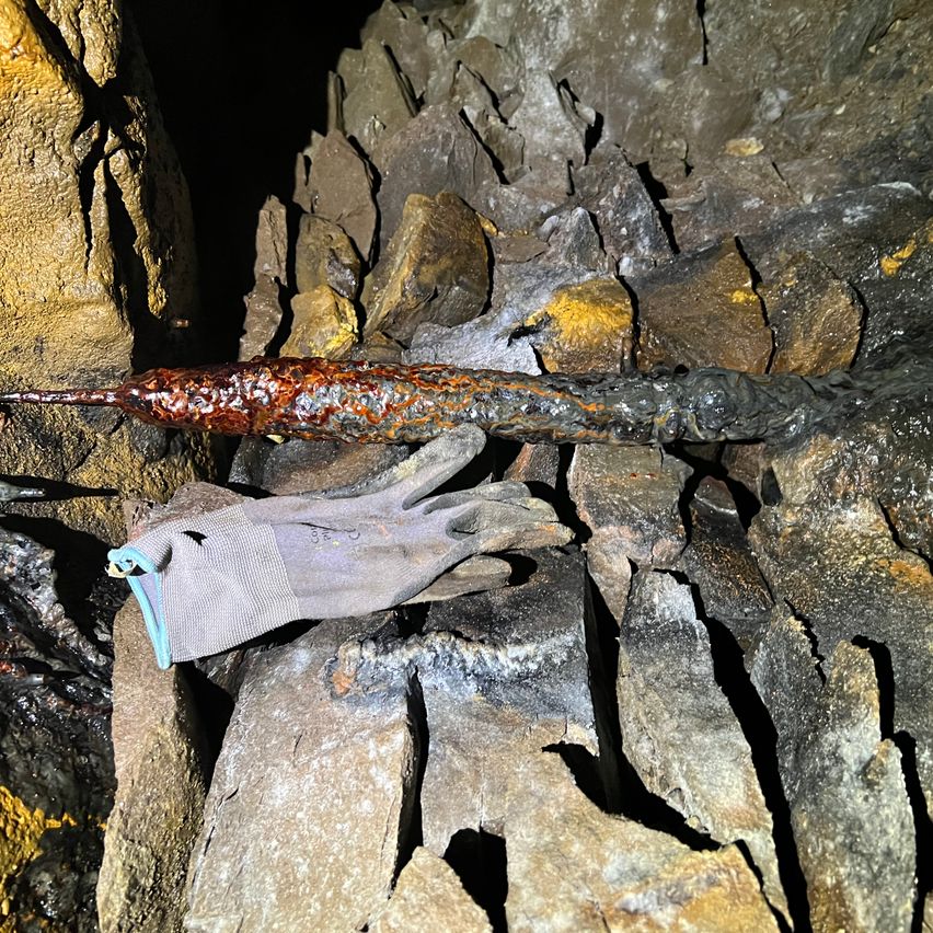 Ein verrostetes Metallrohr mit einem grauen Handschuh auf einer felsigen Oberfläche, in einem dunklen Bereich.
