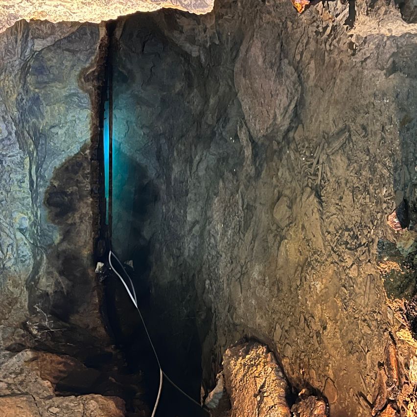 Ein schmaler Durchgang in einer Höhle führt zu einem hellblauen Licht. Die Wände sind rau und uneben, einige Teile zeigen Anzeichen von Erosion.