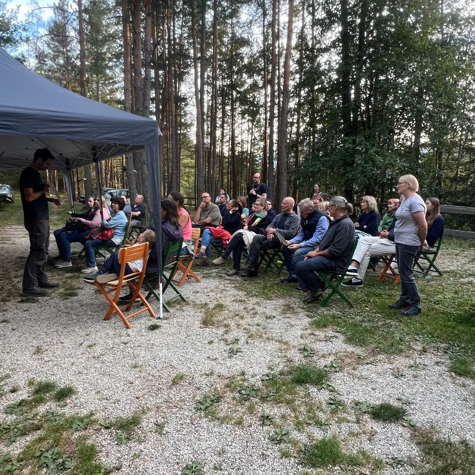 Eine Gruppe von Menschen ist in einem Kreis unter einem Zelt im Wald versammelt.