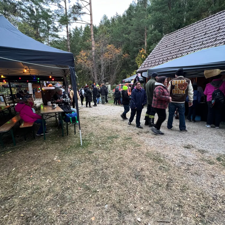 Eine Gruppe von Menschen versammelt sich bei einer Outdoor-Veranstaltung in einem bewaldeten Gebiet. Einige sitzen an Tischen unter blauen Zelten, während andere herumlaufen. Die Umgebung ist von Bäumen umgeben und es sind zwei Zelte zu sehen.