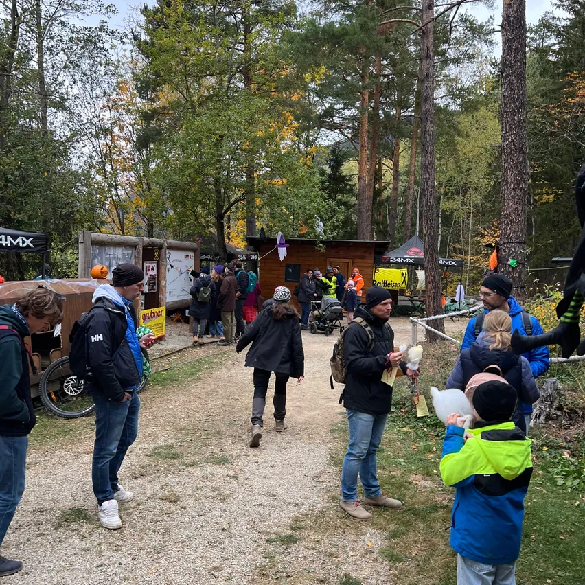 Eine Gruppe von Menschen steht und geht auf einem Kiesweg im Wald. Einige tragen Rucksäcke und Mützen. Im Hintergrund befinden sich kleine Holzhütten mit Schildern und Kürbissen.