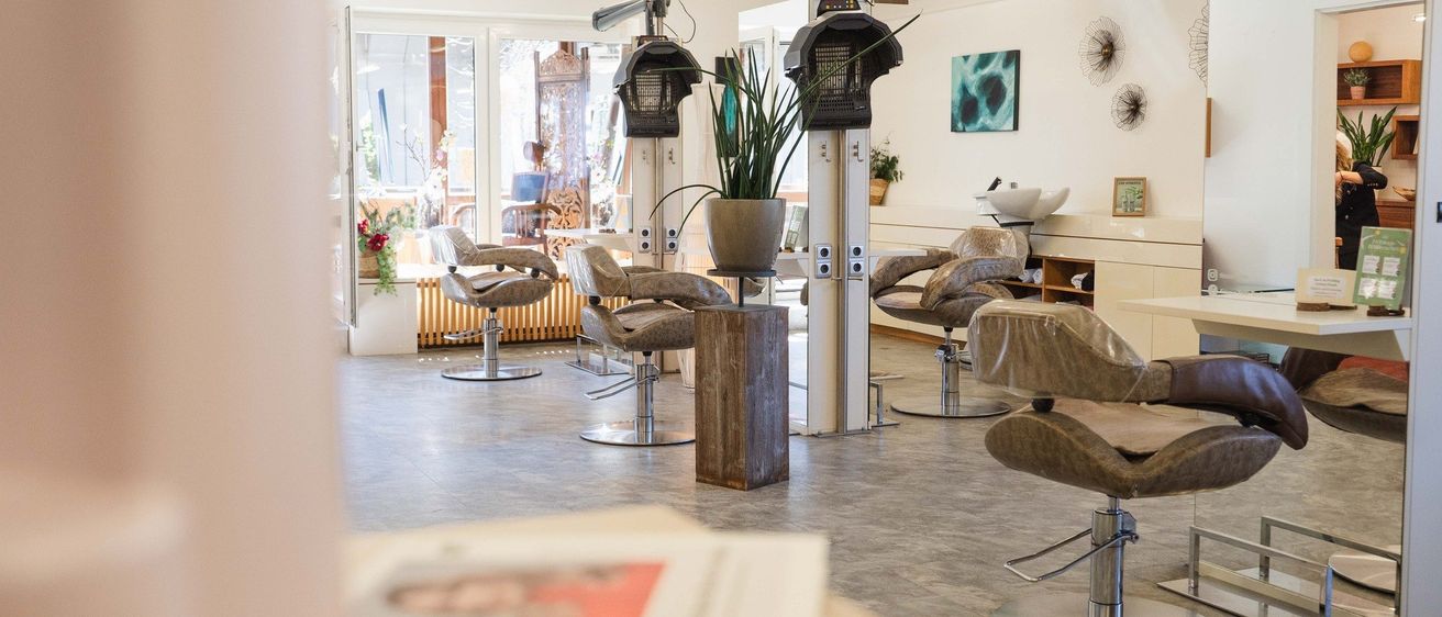 Ein moderner Salon mit weißen Wänden und grauen Böden. Er verfügt über mehrere Stühle und einen Holzhocker mit einer Pflanze. Der Raum ist mit Deckenleuchten gut beleuchtet.