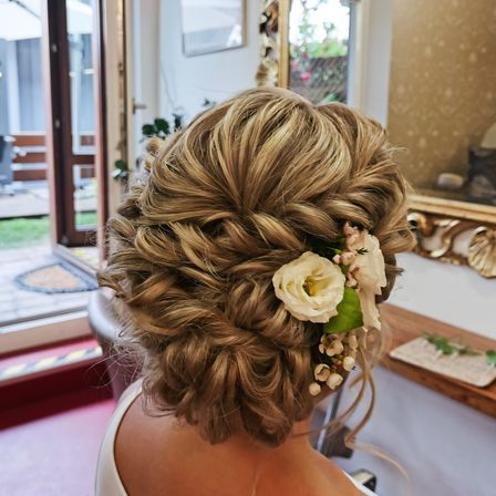 Eine Frau mit einer eleganten Frisur, geschmückt mit einer weißen Blume und Grünpflanzen, sitzt in einem Salon, mit einem goldenen Spiegel und einer offenen Glastür im Hintergrund.