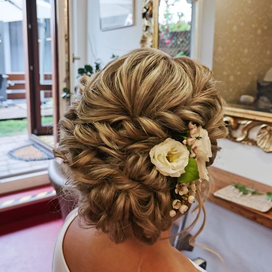 Eine Frau mit einer eleganten Frisur, geschmückt mit einer weißen Blume und Grünpflanzen, sitzt in einem Salon, mit einem goldenen Spiegel und einer offenen Glastür im Hintergrund.