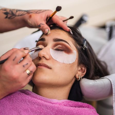 Eine Frau mit einem rosa Bademantel sitzt, während eine Visagistin ihr Make-up aufträgt, mit Pinseln und weißen Pflastern über ihren Augen.