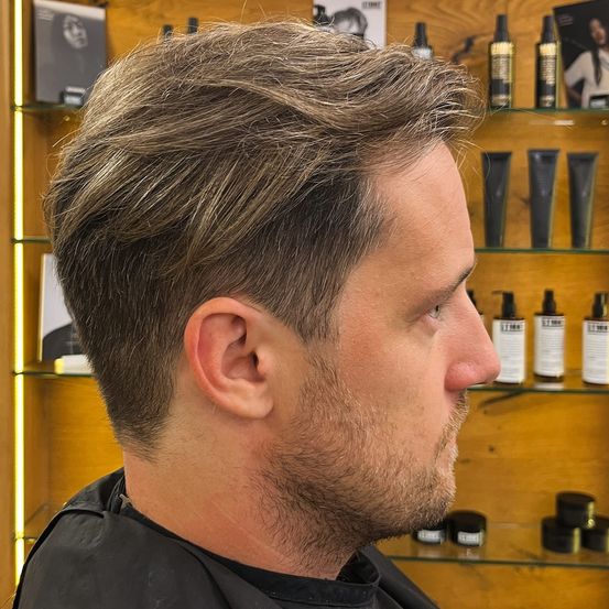 Ein Mann mit kurzen Haaren und Bart steht vor einem Friseurarbeitsplatz. Der Mann trägt eine schwarze Schürze. Dahinter befinden sich Regale mit Haarpflegeprodukten.