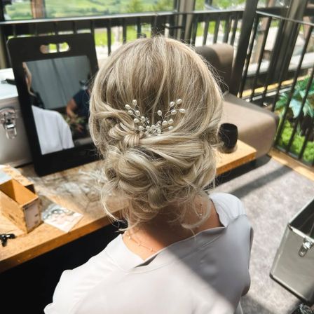 Eine Frau mit blondem Haar, geschmückt mit einer Perlenhaarnadel, sitzt an einem Schreibtisch. Hinter ihr spiegelt ein Spiegel die Aussicht auf das Freie. Ein Sofa, ein Tisch und dekorative Gegenstände sind im Raum zu sehen.