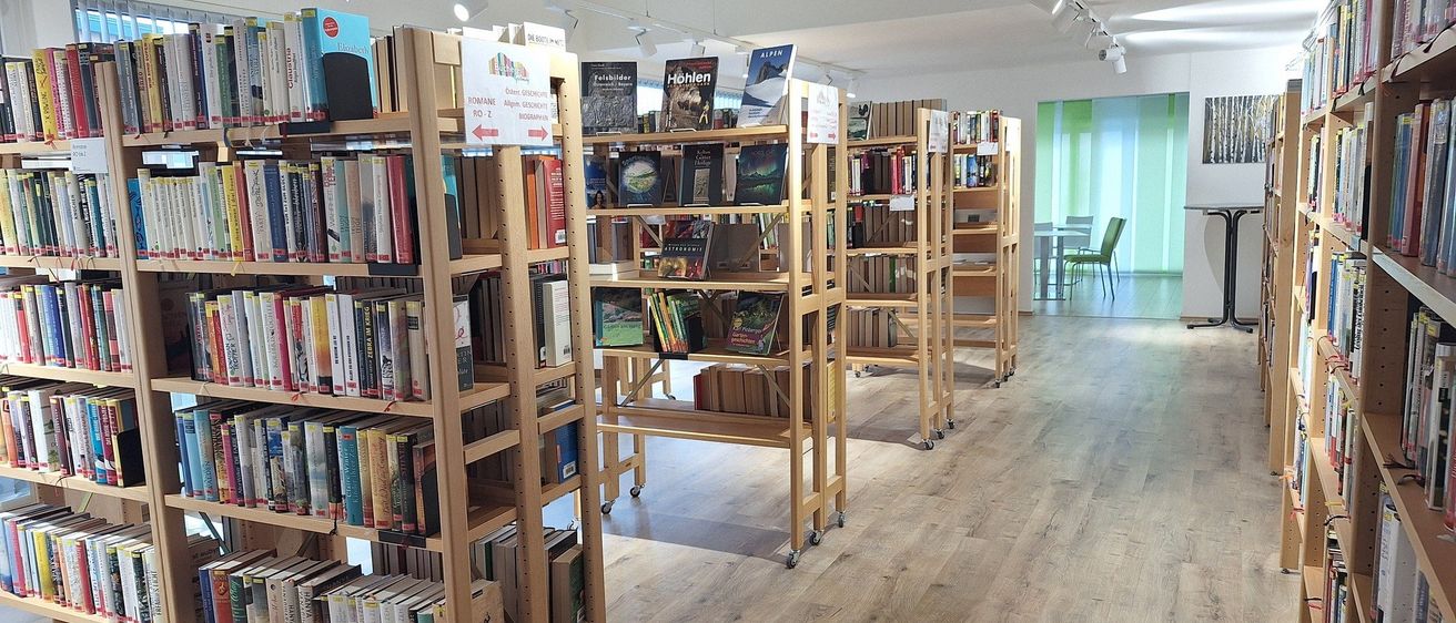 Eine Innenbibliothek mit Holzregalen voller Bücher. Der Raum hat Holzböden und weiße Wände. Es hängen Lampen an der Decke.