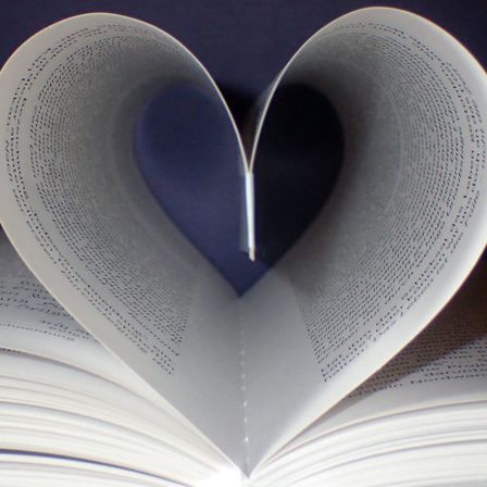 Ein offenes Buch ist in Form eines Herzens gefaltet und zeigt einen blauen Hintergrund hinter den Seiten. Die Seiten sind mit Text gefüllt und das Buch ist leicht an den Seiten angehoben.