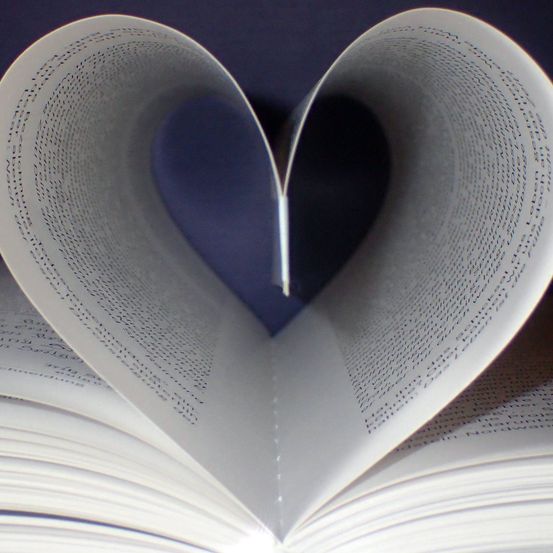 Ein offenes Buch ist in Form eines Herzens gefaltet und zeigt einen blauen Hintergrund hinter den Seiten. Die Seiten sind mit Text gefüllt und das Buch ist leicht an den Seiten angehoben.