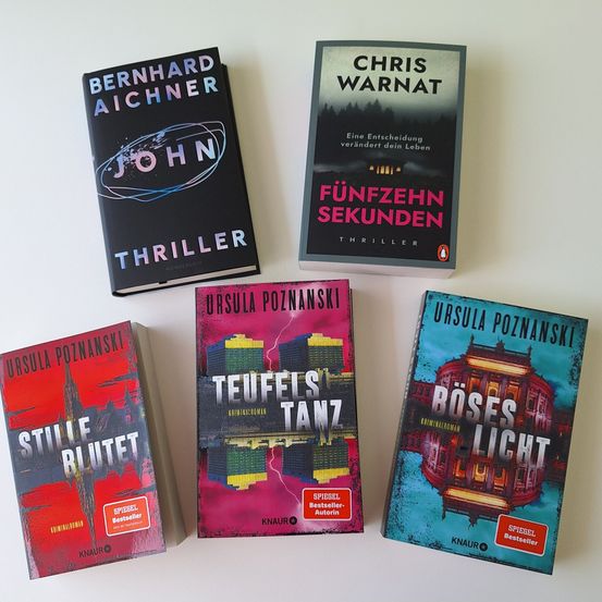 Sechs Bücher auf einer weißen Oberfläche angeordnet. Jedes Buchcover zeigt unterschiedliche Designs und Titel. Von links nach rechts sind die Titel 'John', 'Fünfzehn Sekunden', 'Teufelstanz', 'Stille Blutet' und 'Böses Licht'. Die Bücher sind hauptsächlich in Schwarz und Rot gehalten, mit einigen Elementen in Weiß.