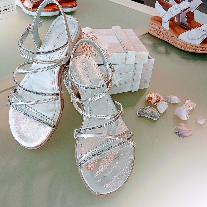 Ein Paar silberne Sandalen mit Kristallverzierungen werden auf einem Glastisch mit dekorativen Muscheln ausgestellt.