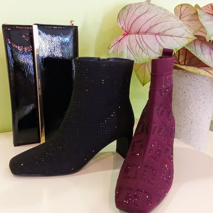 Ein Paar schwarze und lila hochhackige Stiefel mit Glitzer und eine schwarze Clutch-Tasche sind auf einer weißen Oberfläche ausgestellt.