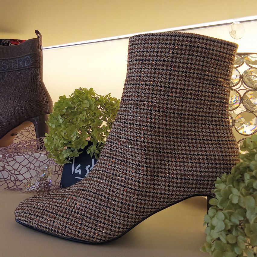 Ein brauner karierter Stiefel mit hohem Absatz, ausgestellt auf einer weißen Oberfläche mit einer Pflanze und einem anderen Schuh im Hintergrund.