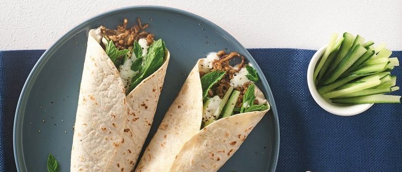 Zwei Lamm- und Linsen-Wraps mit Gurke und Minze auf einem blauen Teller, begleitet von einer kleinen Schüssel mit geschnittenen Gurken.