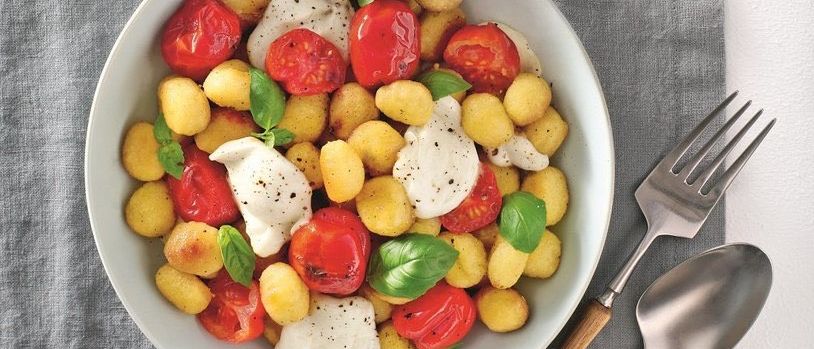 Eine weiße Schüssel mit Kartoffeln, Tomaten und Mozzarella steht auf einem grauen Tuch neben einer Gabel und einem Löffel.