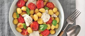 Eine weiße Schüssel mit Kartoffeln, Tomaten und Mozzarella steht auf einem grauen Tuch neben einer Gabel und einem Löffel.