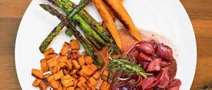 Ein Teller mit Steak, roten Zwiebeln, Karotten und Spargel. Das Steak ist mit Rosmarin und Rotweinsauce überzogen. Daneben liegen geröstete Süßkartoffeln und ein Holztisch.