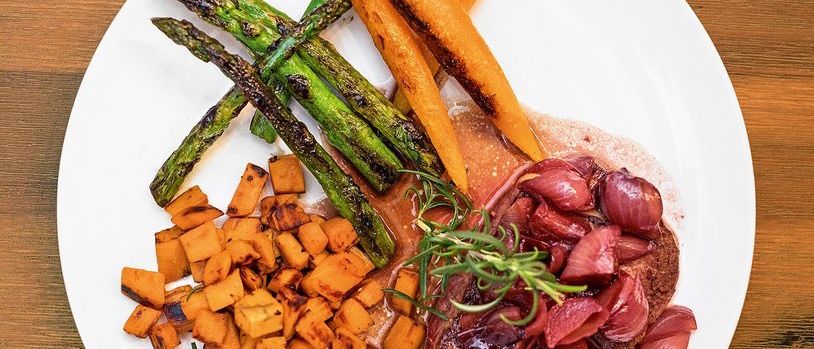 Ein Teller mit Steak, roten Zwiebeln, Karotten und Spargel. Das Steak ist mit Rosmarin und Rotweinsauce überzogen. Daneben liegen geröstete Süßkartoffeln und ein Holztisch.