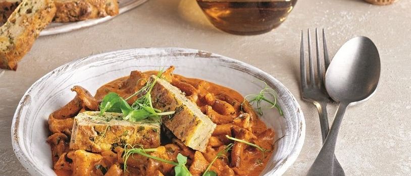 Eine Schüssel mit Pilz- und Bohnencurry mit einem Stück Brot auf einem Teller, ein Glas Wein und ein Löffel und eine Gabel auf einem Tisch.