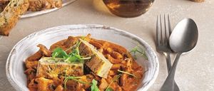 Eine Schüssel mit Pilz- und Bohnencurry mit einem Stück Brot auf einem Teller, ein Glas Wein und ein Löffel und eine Gabel auf einem Tisch.