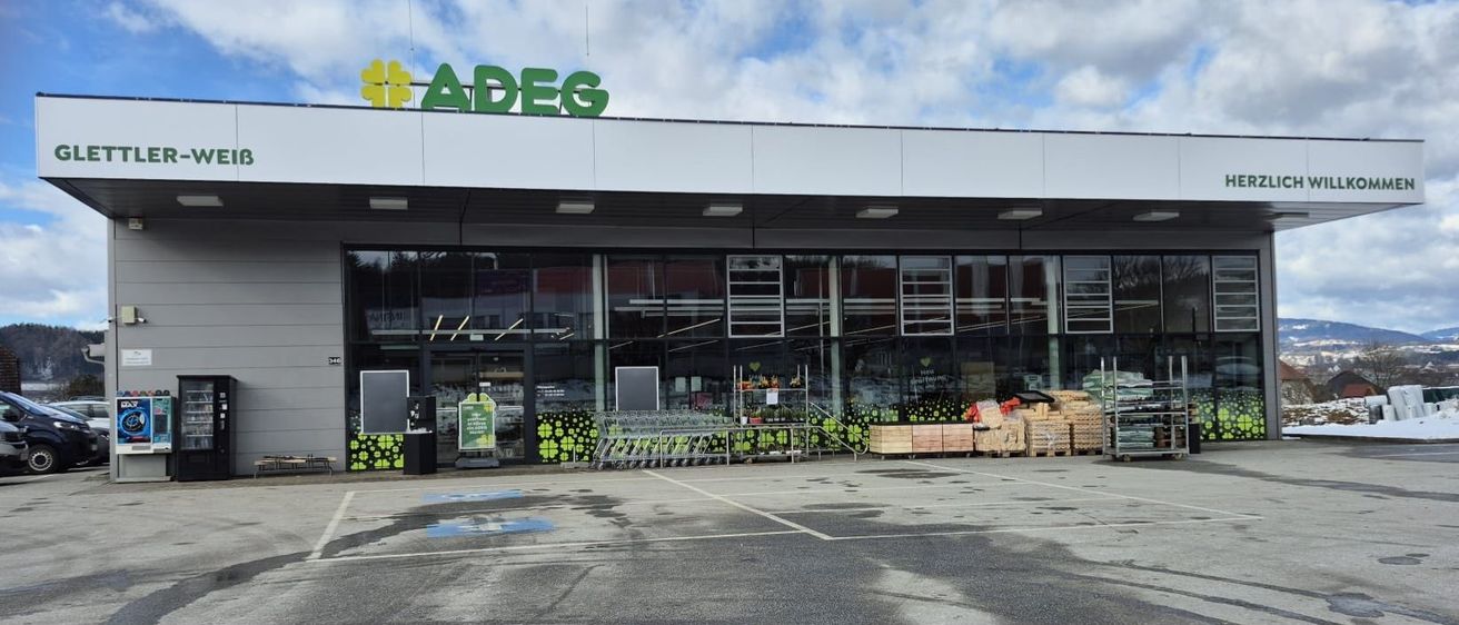 Die Vorderseite eines ADEG-Geschäfts, ein grünes und gelbes Schild oben. Der Parkplatz ist leer, mit Trolleys und Topfpflanzen draußen.