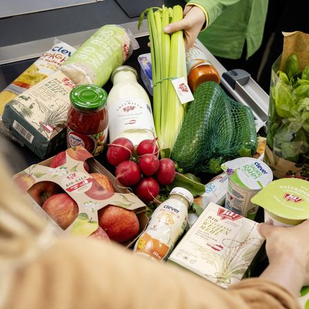 Eine Person in einem grünen Shirt platziert verschiedene Lebensmittel auf einem Kassenband, darunter Sellerie, Radieschen, Äpfel, Joghurt und andere Produkte.