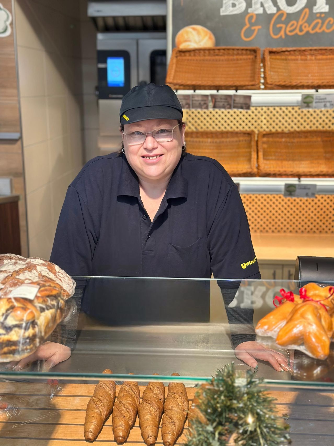 Eine Frau in einer schwarzen Uniform steht hinter einer Glastheke in einer Bäckerei, lächelt und hält ein großes Brot.