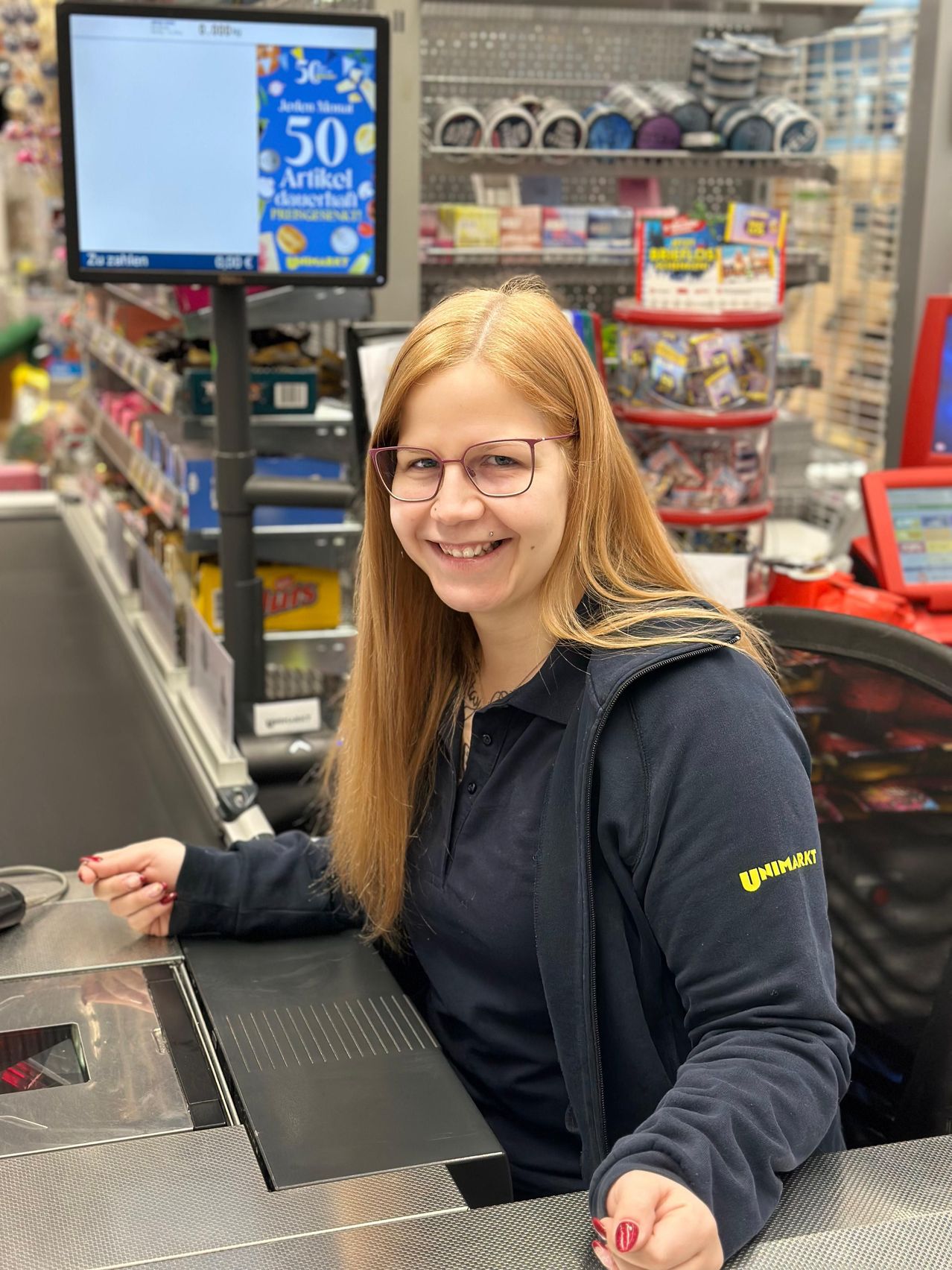 Eine lächelnde Frau mit Brille und einer schwarzen Jacke mit dem Wort Unimarkt sitzt an einer Kasse in einem Geschäft.