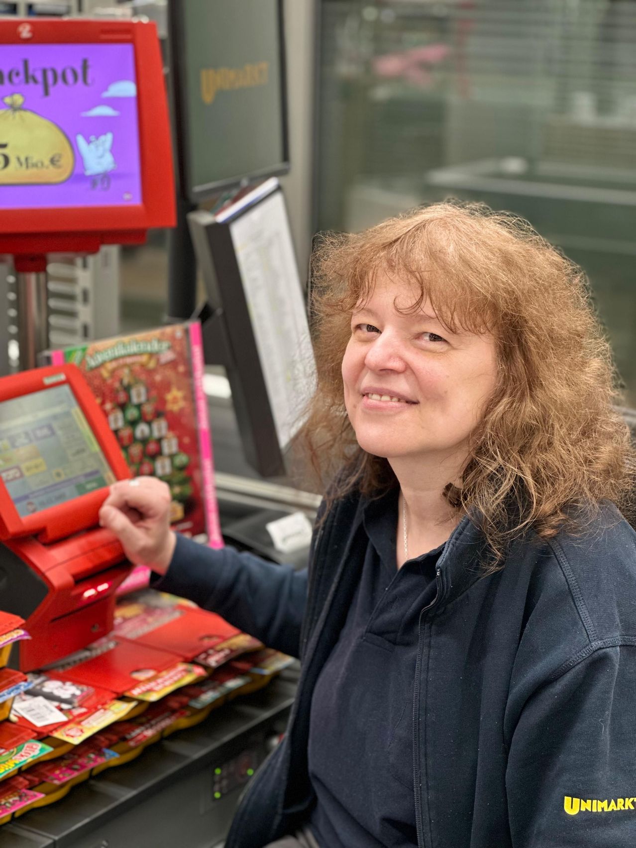 Eine Frau mit lockigen Haaren lächelt und steht hinter einer Kasse. Vor ihr steht ein rotes Gerät mit einem Bildschirm, auf dem 5 Millionen Euro angezeigt werden.