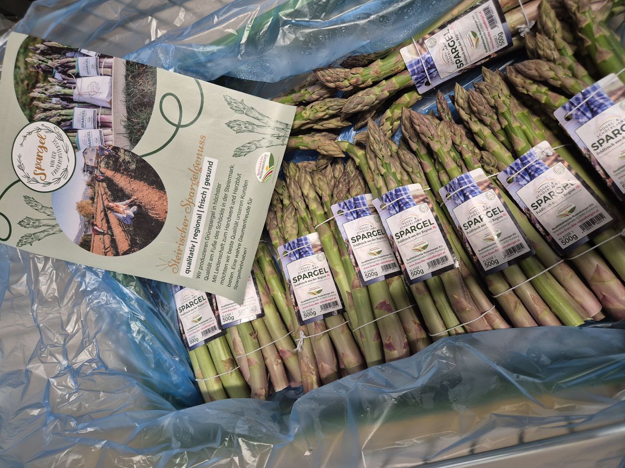 Bündel frischen Spargels in Plastik verpackt, mit Etiketten, die auf biologische und regionale Herkunft hinweisen. Jeder Bund ist mit weißem Faden gebunden und hat ein Preisschild.