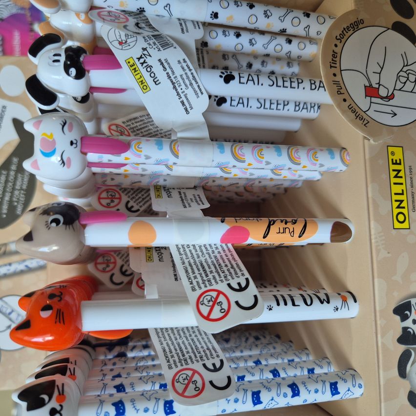 Verschiedene tierförmige Stifte sind in einem Karton gestapelt. Sie haben tierthematische Designs und enthalten Phrasen wie 'Fressen, Schlafen, Bellen'. Jeder Stift hat einen Aufkleber mit Informationen über sein Design.