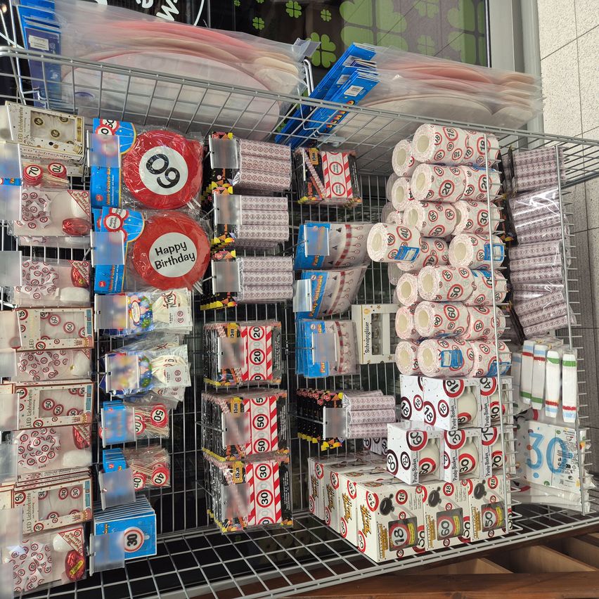 A store shelf displays numerous items, including birthday balloons with numbers 9 and 30, wrapped candies, and various party supplies.