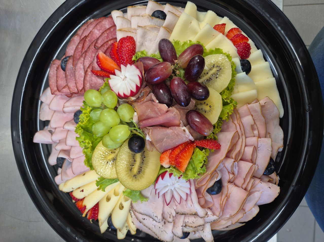 Ein schwarzer Teller mit einer Auswahl an Fleisch, Käse, Trauben, Erdbeeren und Kiwis in einer dekorativen Anordnung.