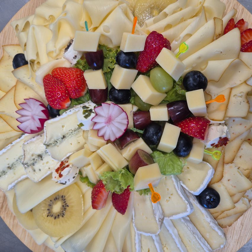 Ein Holztablett mit verschiedenen Käsesorten, Erdbeeren, Trauben und Kiwi, kunstvoll arrangiert. Einige Käsescheiben haben Zahnstocher.