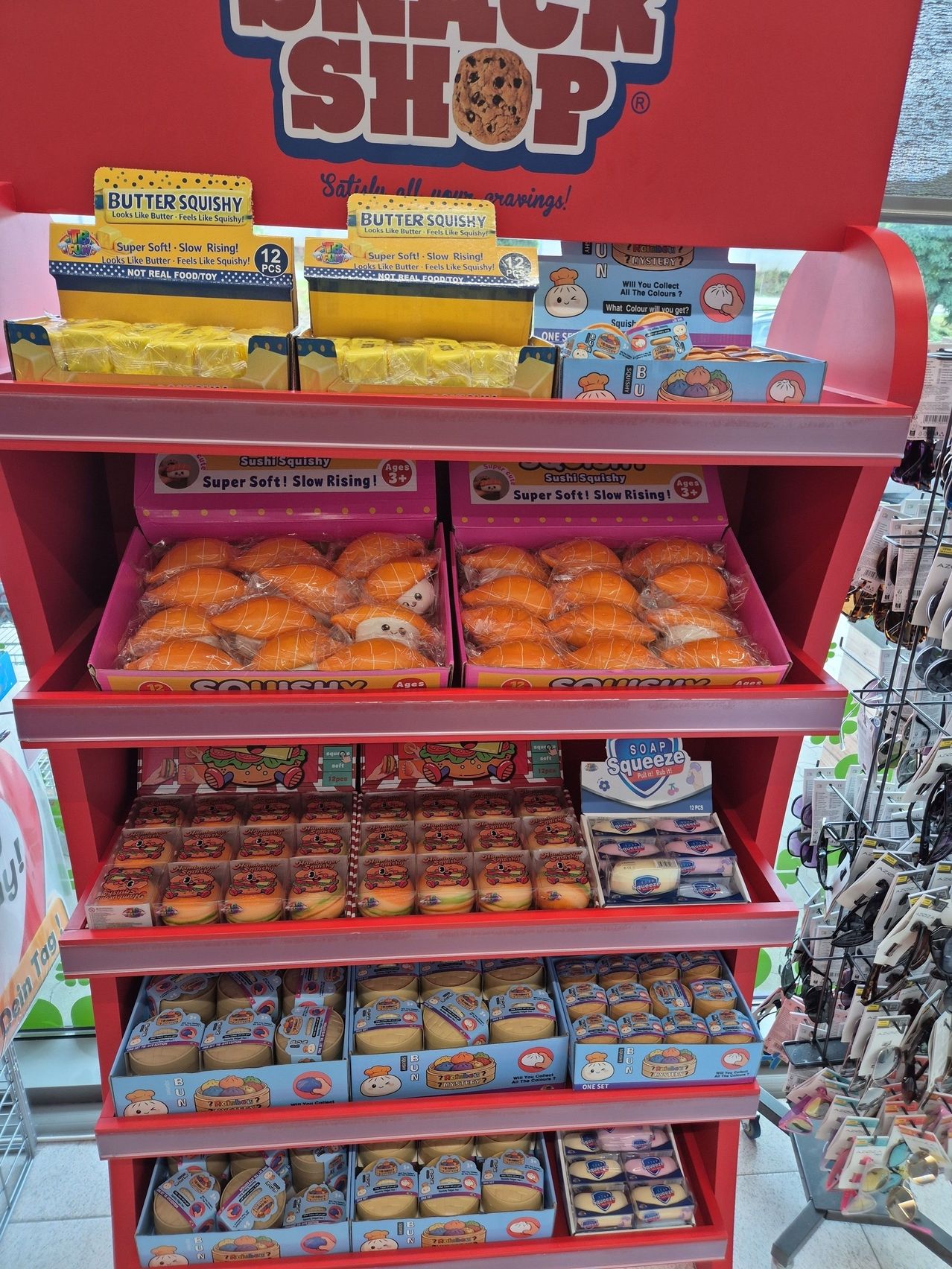 Ein roter Regal zeigt verschiedene Schachteln von Butter Squishy und Sushi Squishy Spielzeug, mit bunten Verpackungen und Charakteren. Die Spielzeuge sind ordentlich in Reihen angeordnet, und die Regale sind mit anderen Produkten gefüllt.